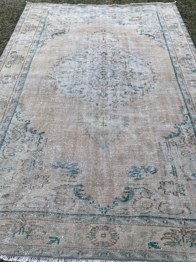 May include: A vintage rug with a faded beige background and a teal floral pattern. The rug has a distressed look and is made of wool.