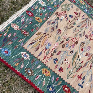 May include: A green and beige rug with a floral pattern. The rug has a fringed border and is made of wool.
