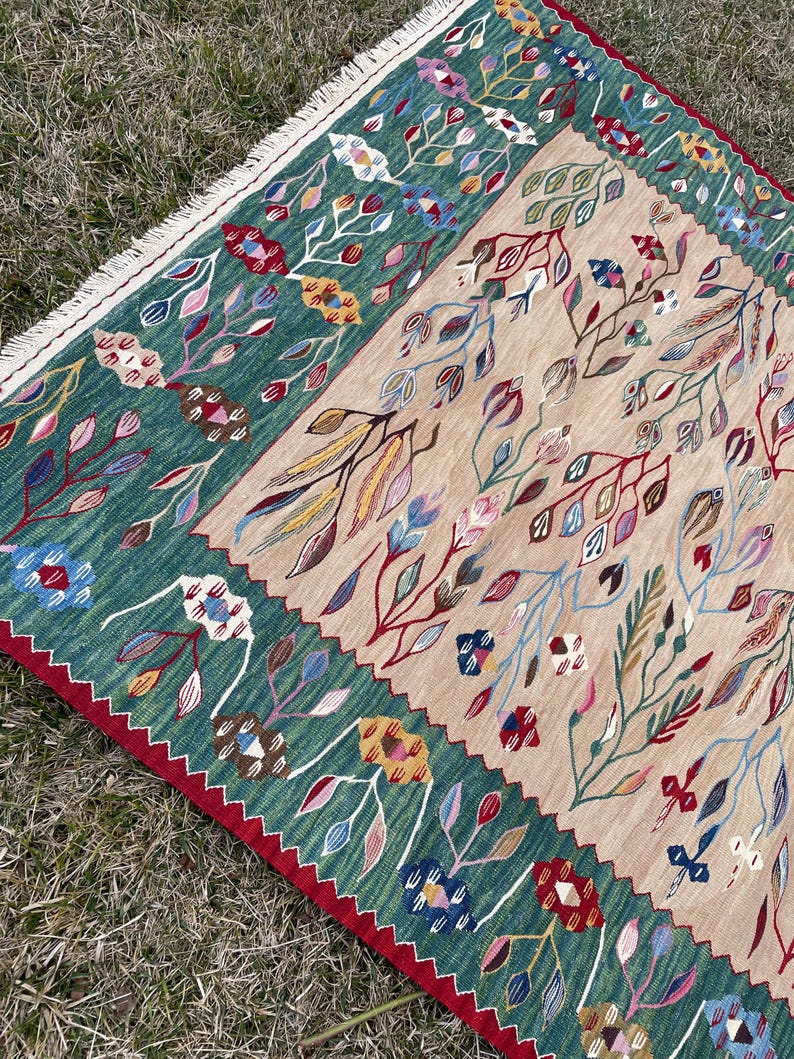 May include: A green and beige rug with a floral pattern. The rug has a fringed border and is made of wool.