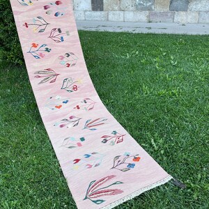 May include: A pink runner rug with a floral design featuring colorful flowers and leaves. The rug has a fringe border.