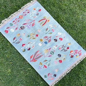 May include: A light blue rug with a white fringe border features a repeating pattern of stylized floral designs in shades of red, pink, green, yellow, and blue.