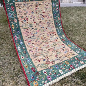 May include: A traditional rug with a green, beige and red colour scheme. The rug features a floral pattern with intricate details. The rug is made of wool and has a fringe border.