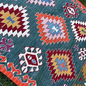 May include: A teal blue rug with a geometric pattern in shades of red, orange, yellow, white, pink, and blue. The rug has a fringed border.