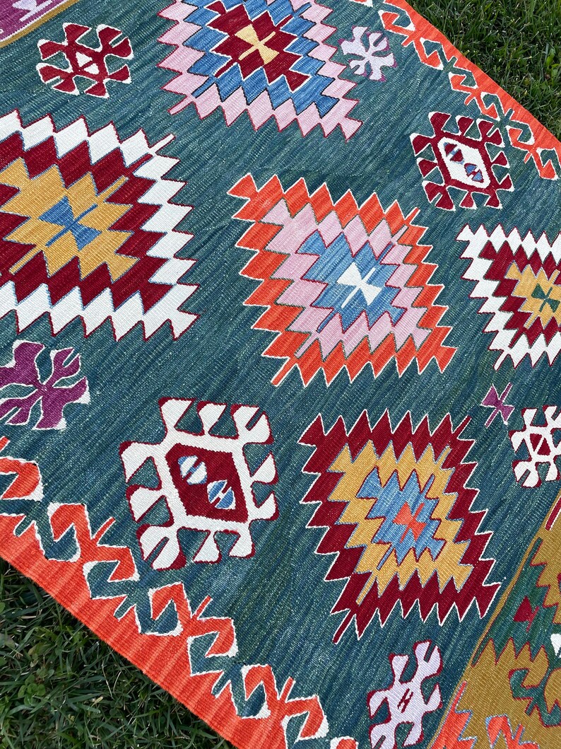 May include: A teal blue rug with a geometric pattern in shades of red, orange, yellow, white, pink, and blue. The rug has a fringed border.