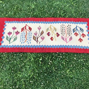 May include: A red, white, and blue patterned rug with a floral design. The rug has a fringe on both ends.