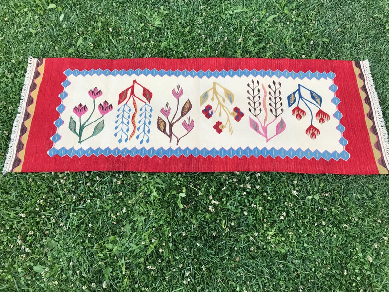 May include: A red, white, and blue patterned rug with a floral design. The rug has a fringe on both ends.