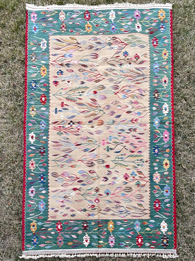 May include: A colourful rug with a green border and a beige centre featuring a floral pattern. The rug has a fringe border.