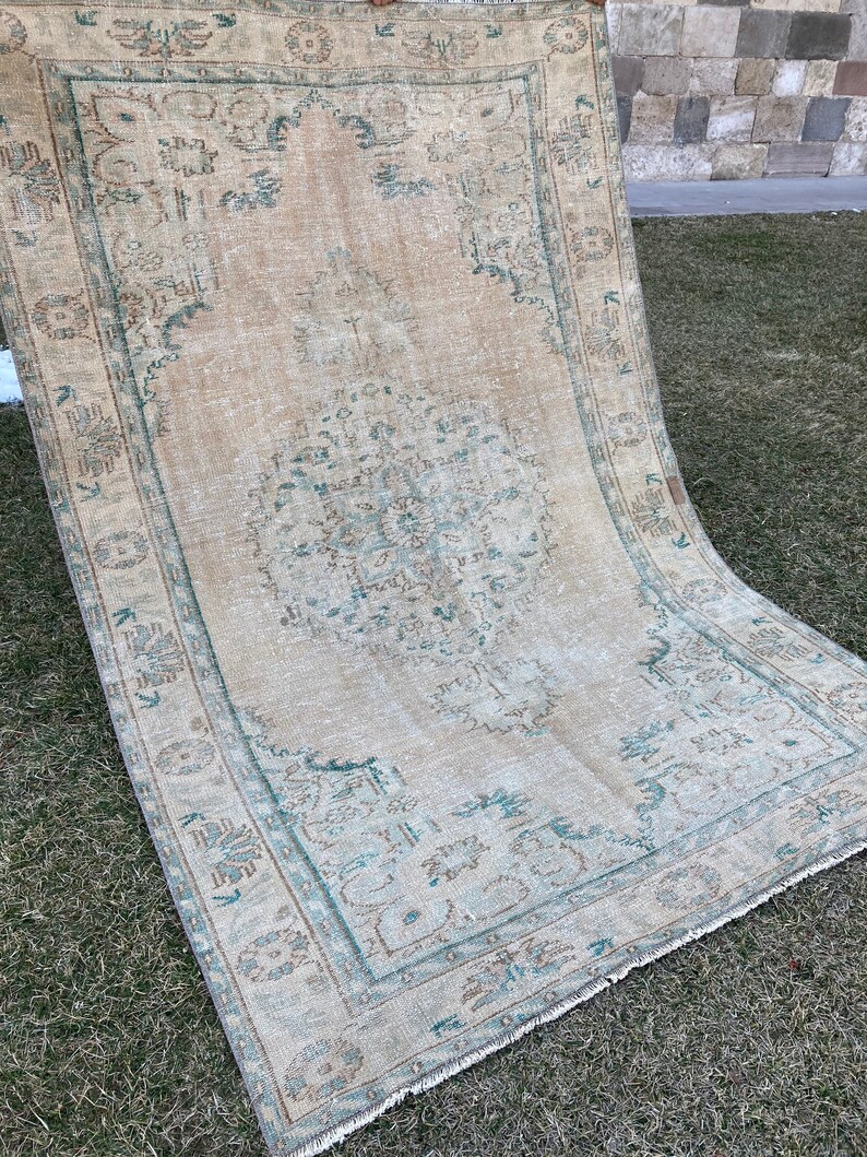 May include: A vintage rug with a light beige background and a teal blue pattern. The rug has a faded look and a fringed edge.