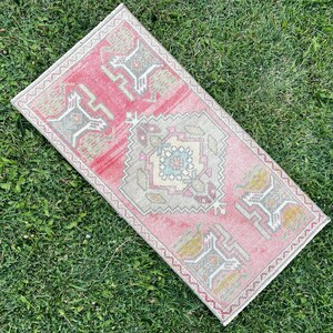 May include: A rectangular, vintage-style rug with a pink and beige color scheme. The rug features a central geometric design with a border of repeating patterns. The rug is placed on a bed of green grass.
