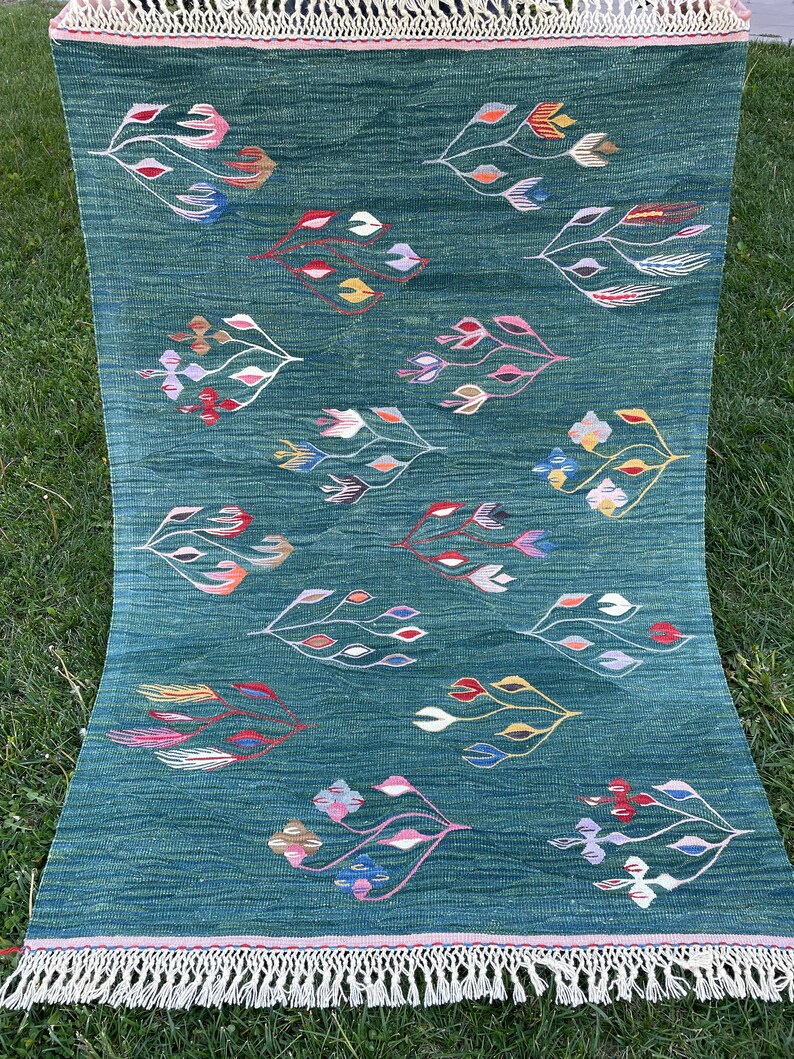 May include: A green rug with a white fringe border features a repeating pattern of stylized floral designs in shades of pink, blue, yellow, and red.