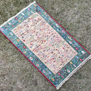 May include: A rectangular rug with a cream background and a green border. The rug features a floral pattern with small, colourful flowers and leaves.