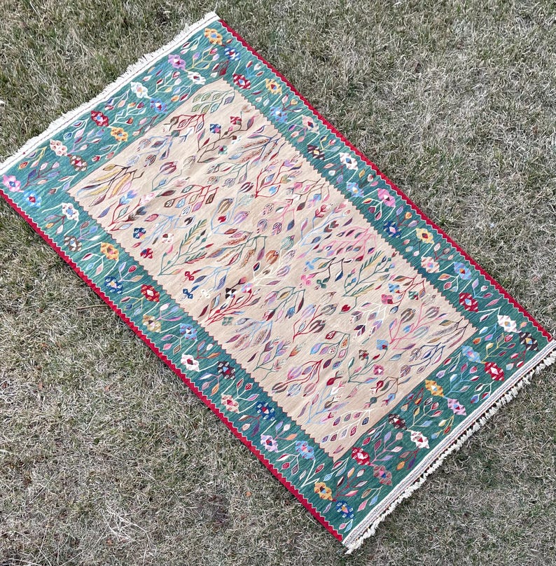May include: A rectangular rug with a cream background and a green border. The rug features a floral pattern with small, colourful flowers and leaves.