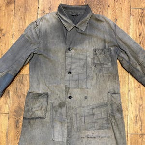 May include: A long-sleeved, button-down coat in a muted, weathered gray tone. The garment features a pointed collar, a single chest pocket, two large lower pockets, and visible stitched patchwork detailing throughout the fabric surface.