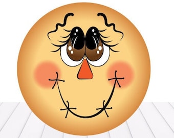 May include: A round, yellow scarecrow face with stitched-on eyes, a nose, and a mouth. The face has rosy cheeks and long eyelashes.