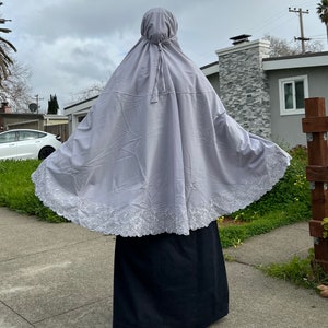 May include: A light gray prayer garment with a hood and a decorative white lace trim along the bottom edge. The garment is worn over a black skirt. The image is taken outdoors on a cloudy day.