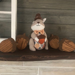 May include: A plush squirrel wearing a brown top hat and a bow tie holds an acorn. The squirrel is sitting on a wooden shelf with four wooden acorns on either side.