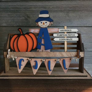 May include: A wooden fall-themed decoration with a scarecrow, pumpkin, and a sign that reads "Hayrides, Pumpkin Patch, Corn Maze". The decoration also features a banner that says "Fall" in blue letters.