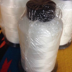 May include: Three cylindrical spools of white thread, each wrapped in clear plastic. The spools have black caps and sit on a dark base. The thread appears to be tightly wound. The background is a colorful textile.