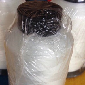 May include: Three white spools of thread, each wrapped in clear plastic. The spools have black caps. The background is a blurred mix of colors. The image is well-lit, highlighting the texture of the thread.