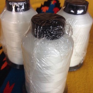 May include: Three spools of white thread wrapped in clear plastic, each with a black lid. The spools are sitting on a colorful patterned surface.