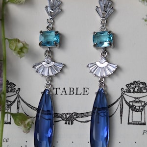 Peut inclure: Une paire de boucles d'oreilles pendantes en argent avec des pierres précieuses bleues et turquoise. Les boucles d'oreilles présentent une pierre précieuse bleue en forme de larme, une pierre précieuse turquoise rectangulaire et un motif en éventail avec des strass clairs.