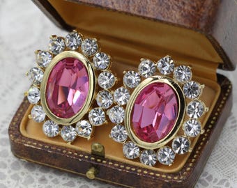 May include: A pair of vintage clip-on earrings with large pink oval center stones surrounded by clear crystals. The earrings have a gold-tone setting and are displayed in a brown leather jewelry box with a gold interior.