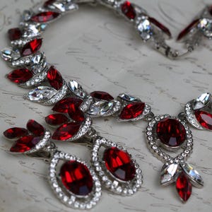 Peut inclure: Un collier et des boucles d'oreilles argentés ornés de cristaux rouges et transparents. Le collier présente une pierre rouge ovale centrale et un motif en forme de feuille. Les boucles d'oreilles sont en forme de goutte avec des pierres rouges.