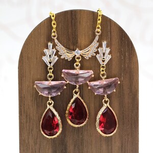May include: A gold-toned necklace with a crescent-shaped top adorned with clear crystals. The necklace features three pendants: two half-moon shaped pink gems and three teardrop-shaped red gems. The necklace is displayed on a wooden stand.