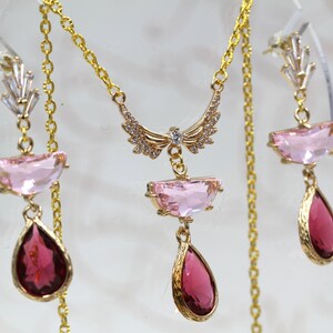 May include: A gold-toned jewelry set featuring a necklace and earrings. The necklace has a pendant with a wing-like design and a pink rectangular stone. The earrings have pink and red teardrop-shaped stones.