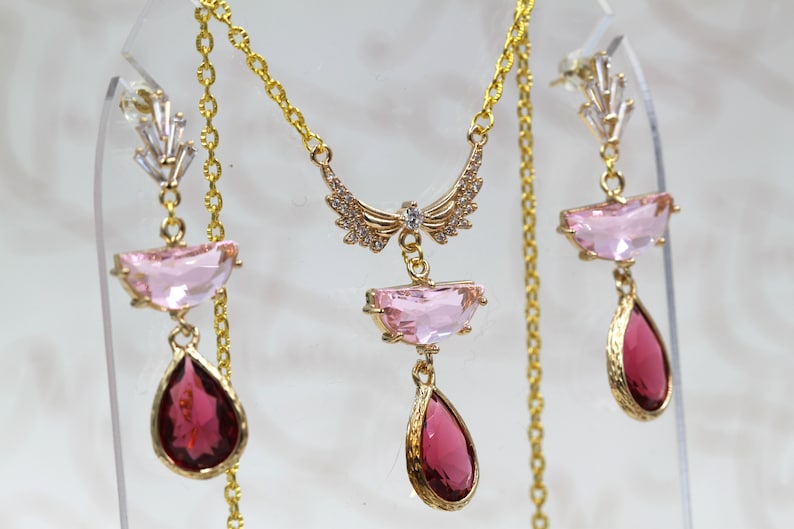 May include: A gold-toned jewelry set featuring a necklace and earrings. The necklace has a pendant with a wing-like design and a pink rectangular stone. The earrings have pink and red teardrop-shaped stones.