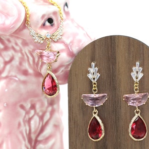 May include: A gold-toned necklace and earrings set. The necklace features a pendant with a pink and red gemstone design. The earrings have a similar design, with pink and red gemstones. The jewelry is displayed on a wooden stand and a pink ceramic dog.