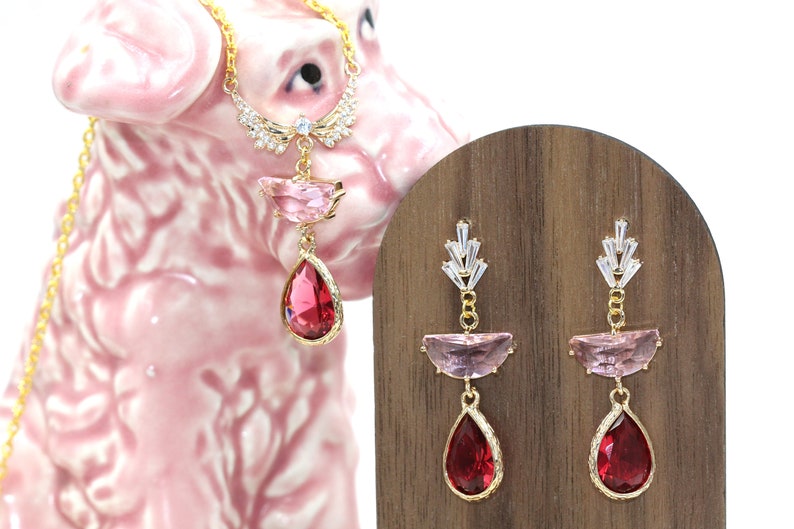 May include: A gold-toned necklace and earrings set. The necklace features a pendant with a pink and red gemstone design. The earrings have a similar design, with pink and red gemstones. The jewelry is displayed on a wooden stand and a pink ceramic dog.