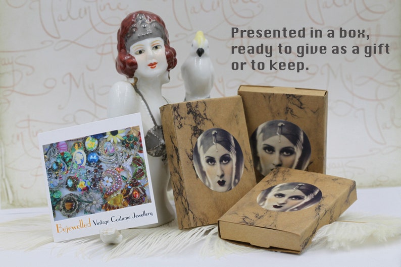 May include: An arrangement of vintage costume jewelry, presented in boxes with a vintage portrait. The boxes are brown with a marble design. A card displays various colorful brooches and jewelry. Text reads "Bejewelled Vintage Costume Jewellery."