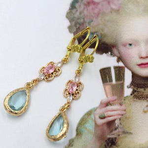 Puede incluir: Pendientes colgantes dorados con una piedra en forma de lágrima azul claro. Los pendientes presentan piedras preciosas rosas y blancas y detalles florales. El fondo incluye una ilustración vintage de una mujer.