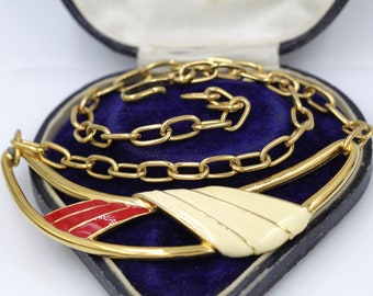 Puede incluir: Collar dorado con diseño de esmalte crema y rojo, presentado en una caja de joyería negra con interior de terciopelo azul. El collar incluye una cadena y un colgante decorativo.