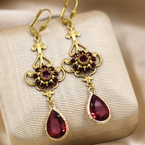 Peut inclure: Une paire de boucles d'oreilles en or avec des cristaux rouges en forme de goutte qui pendent. Les boucles d'oreilles présentent un motif floral avec de plus petits cristaux rouges.
