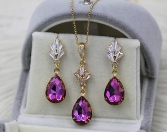 Puede incluir: Un conjunto de joyas dorado que incluye un collar y pendientes a juego. El conjunto incluye un colgante y dos pendientes en forma de lágrima con una vibrante piedra preciosa magenta. Los pendientes y el colgante están acentuados con cristales transparentes con corte de baguette.