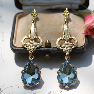 May include: A pair of gold-tone dangle earrings with a teardrop-shaped blue crystal. The earrings feature a floral design with a delicate filigree pattern.