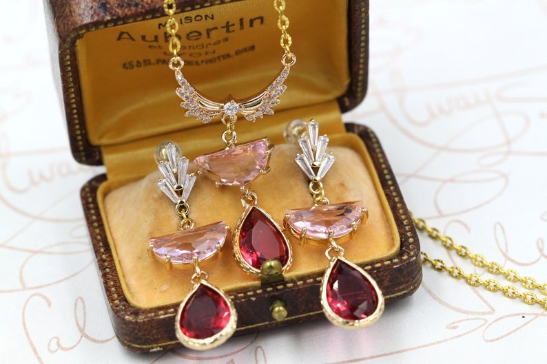 May include: A gold-toned necklace and earrings set displayed in a vintage jewelry box. The necklace features a crescent-shaped pendant with clear crystals. The earrings and necklace have pink and red gemstones.