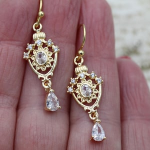 May include: A pair of gold dangle earrings with clear crystal stones. The earrings feature a teardrop-shaped stone hanging from a delicate, ornate design.
