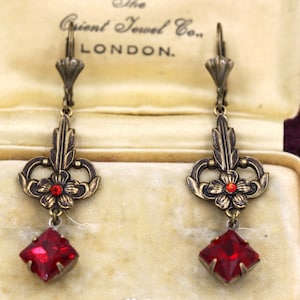 Könnte beinhalten: Ein Paar antike Hängeohrringe mit einem roten Edelstein und kunstvollen, goldfarbenen Metallarbeiten. Die Ohrringe befinden sich in einer kleinen Schachtel mit dem Text "The Orient Jewel Co. LONDON."