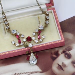May include: Vintage-style necklace with clear and ruby-colored rhinestones set in a gold-tone metal. The necklace is displayed in a pink jewelry box with a cream-colored interior. A vintage photograph is in the background.