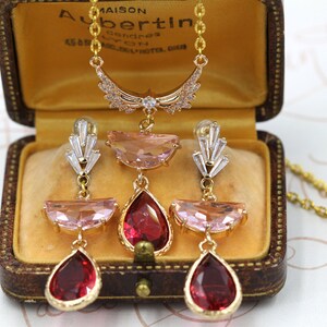 May include: A gold-toned necklace and earring set with pink and red gemstones, displayed in a vintage brown jewelry box. The necklace features a crescent-shaped pendant with clear crystals. The earrings have a similar design, with teardrop-shaped red gems.