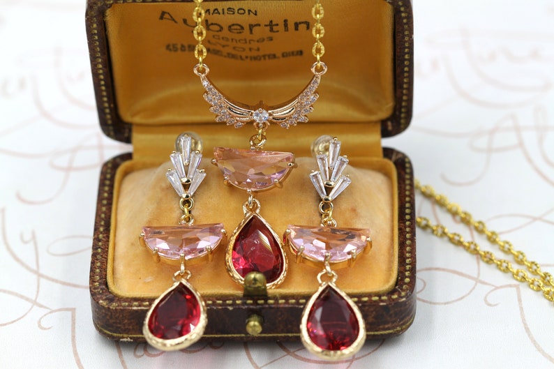 May include: A gold-toned necklace and earring set with pink and red gemstones, displayed in a vintage brown jewelry box. The necklace features a crescent-shaped pendant with clear crystals. The earrings have a similar design, with teardrop-shaped red gems.