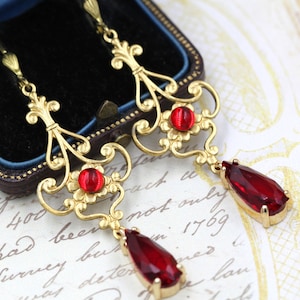 May include: Gold filigree earrings with red teardrop-shaped glass stones. The earrings have a delicate, ornate design.