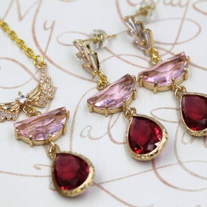 May include: A gold-toned jewelry set featuring a necklace and earrings. The necklace has a delicate chain and a pendant with pink and red gemstones. The earrings have a similar design, with pink and red teardrop-shaped gems. The set is displayed on a patterned surface.