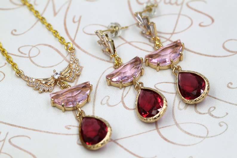 May include: A gold-toned jewelry set featuring a necklace and earrings. The necklace has a delicate chain and a pendant with pink and red gemstones. The earrings have a similar design, with pink and red teardrop-shaped gems. The set is displayed on a patterned surface.
