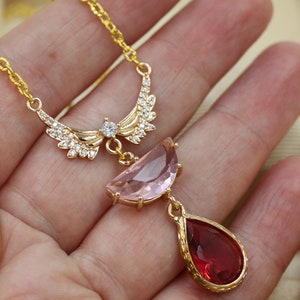 May include: A gold-colored necklace featuring a pendant with a pink, half-moon shaped stone and a red, teardrop-shaped stone. The necklace has a delicate chain and a decorative wing-like design with clear crystals.