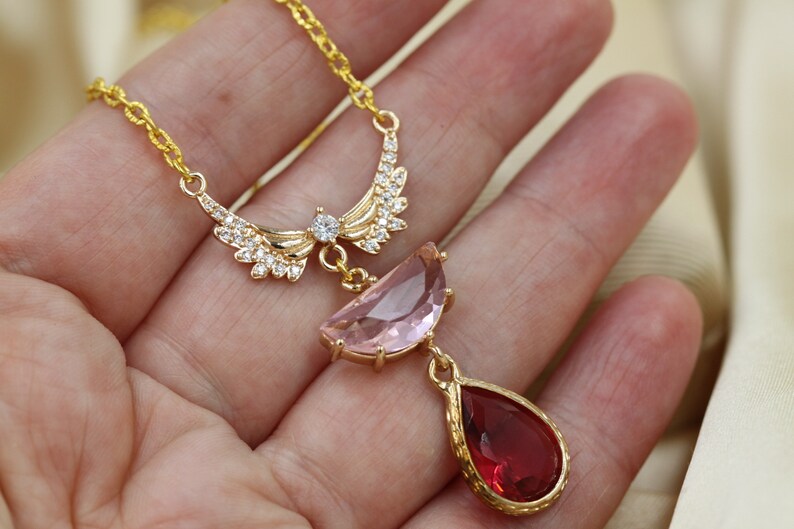 May include: A gold-colored necklace featuring a pendant with a pink, half-moon shaped stone and a red, teardrop-shaped stone. The necklace has a delicate chain and a decorative wing-like design with clear crystals.