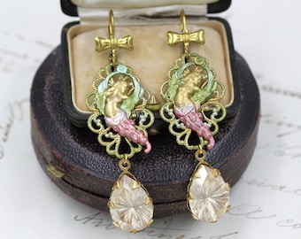 Puede incluir: Un par de pendientes antiguos dorados. Cada pendiente presenta un diseño de filigrana con una figura pintada, detalles en verde y rosa, y un colgante de cristal en forma de lágrima. Los pendientes se exhiben en un joyero vintage.